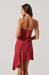 A woman is wearing a sleeveless, red satin slip dress with thin spaghetti straps. The dress features a subtle sheen and a slightly asymmetrical hemline that drapes longer on one side.